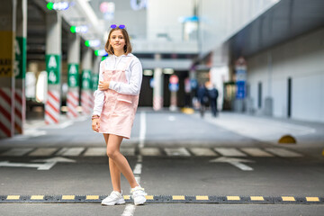 Fototapeta premium young blonde schoolgirl in a pink dress and sunglasses posing in the parking lot