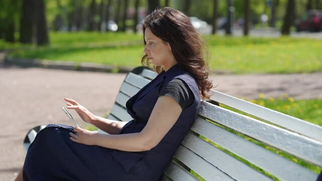 Adult woman using smartphone sitting on bench in a park, brunette woman texting sharing messages on social media.