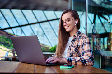 Happy woman freelancer in wireless headphones and eyeglasses using a laptop for remotely working...