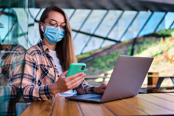 Casual woman freelancer in face mask, wireless headphones and eyeglasses using phone and laptop for remotely working online in cafe. Modern people with mobility lifestyle