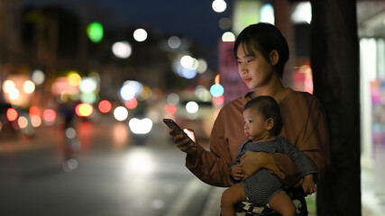 Asian woman and baby wait for her private taxi by using a transportation app on the night street.