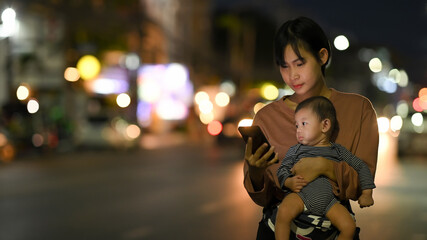 Asian woman and baby wait for her private taxi by using a transportation app on the night street.