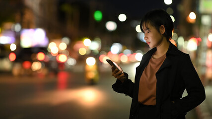 Asian woman waits for her private taxi by using a transportation app on the night street.