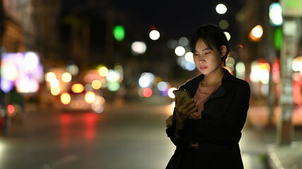 Fototapeta premium Asian woman waits for her private taxi by using a transportation app on the night street.
