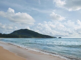 Fototapeta na wymiar Tropical beach with blue sky and island in Thailand. Magnificent seascape, panoramic view. Mountains, sea, sky, clouds. A green hill covered with dense forest on a horizon. Surf, waves, sand, coast. 