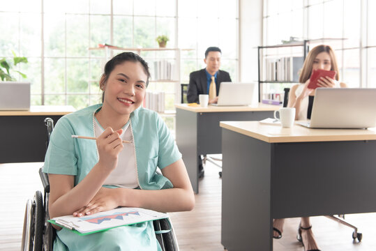 Smiling Asian Woman Office Worker In Wheelchair Holding Pencil Look At Camera, Disabled People Working With Colleague At Workplace