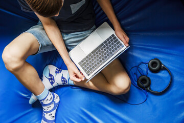 Teen boy sitting with a laptop at home, top view. Learning for a laptop. The guy is sitting on a blue background. Large headphones lie next to the laptop. Home schooling. Distance learning.