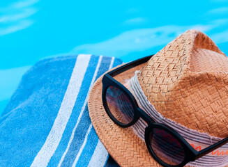 Vintage summer wicker straw beach hat, sun glasses, blue towel near swimming pool, tropical background.