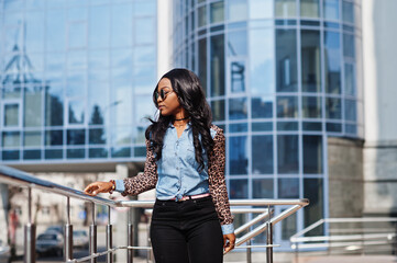Hipster african american girl wearing sunglasses, jeans shirt with leopard sleeves posing at street against modern office building with blue windows.