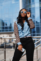 Fototapeta premium Hipster african american girl wearing sunglasses, jeans shirt with leopard sleeves, hold mobile phone, posing at street against modern office building.