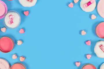 Flat lay with various pink blush beauty products and powder puffs with ribbons and heart shaped pressed powder framing blue background with copy space