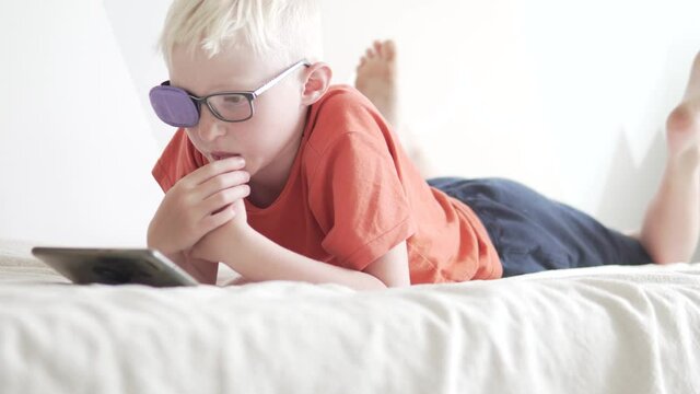 A boy teen with glasses with a taped eye plays in the mobile phone at home.