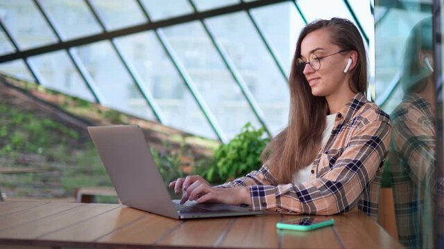 Casual young hipster woman freelancer in wireless headphones and round eyeglasses remotely working online and typing on a laptop in cafe. Modern people with digital smart lifestyle