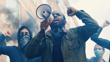 African American young handsome guy with scarf on face protesting in middle of crowd of protesters and screaming mottos in megaphone. Guy leading at manifestation and riot for human rights in smoke. - obrazy, fototapety, plakaty