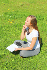 A young beautiful girl in a white T-shirt with her eyes closed sits on the green grass on the lawn with a notebook and a pen, propped up with her hand chin, thinks and dreams, composes text and poems.