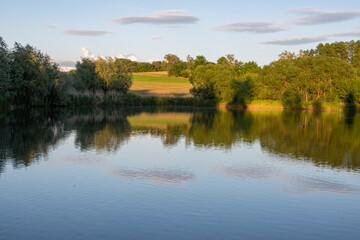 Countryside pond, hills, meadows and forest.  Trees and clouds reflecting into the water. Mirror. Warm sunset light. Country pond. Fishing. Countryside holiday.