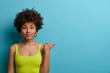 Horizontal shot of calm curly haired Afro American woman points thumb away, wears casual green...