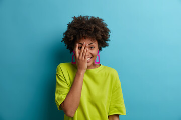 Fototapeta na wymiar Happy teenage girl enjoys moment of fun, covers face with palm, has natural laughter, giggles over something positive, wears green t shirt, isolated on blue background. Good emotions concept