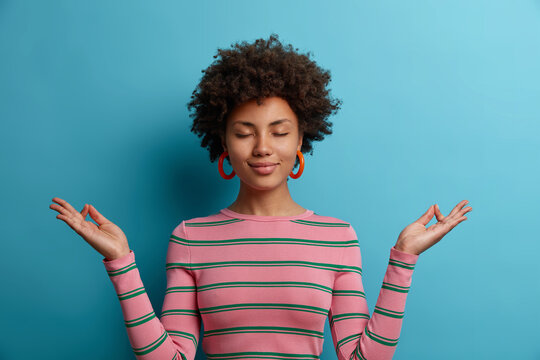 Calm curly haired young woman does meditation gesture, finds harmony in life, closes eyes, stands relieved, has peaceful mind, enjoys yoga practice, poses with calm face against blue background