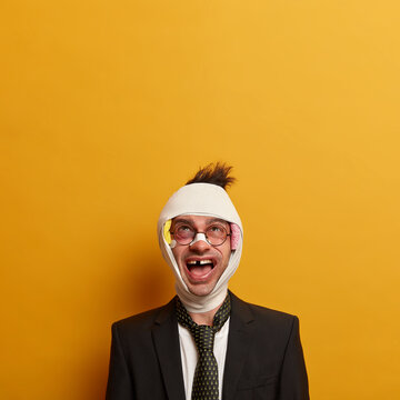 Crazy bruised injured man with plaster on nose, missing teeth, beaten by someone during gang wars, concentrated above, wears tie and black suit, poses against yellow background, copy space upwards