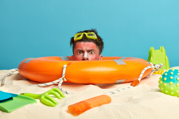Serious man buried in sand, stuck head in lifebuoy, spends free time at beach, poses over blue sea,...