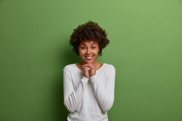 Fototapeta na wymiar Happy positive dark skinned curly woman keeps hands under chin, has pleased optimistic view, wears casual white jumper, isolated on green background, hopes for good, anticipates wonderful news
