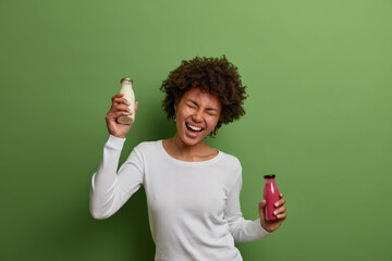 Healthy lifestyle and positive emotions concept. Joyful African American woman dances with bottles of fruit smoothie and plant based milk, has good mood, proper nutriotion, isolated on green wall