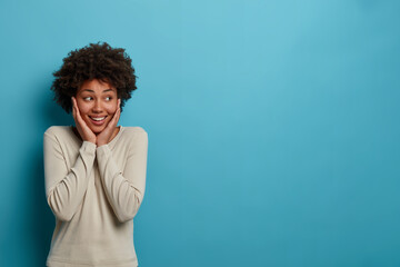 Fototapeta na wymiar So excited! Cheerful pleasant looking dark skinned woman touches cheeks with both hands, looks aside with toothy smile, has positive attitude to life, isolated on blue background, copy space aside
