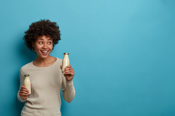 Photo of pleased dark skinned curly girl holds almond milk in glass bottle, finds alternative...