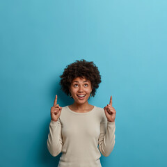 Vertical shot of cheerful woman with Afro haircut points index fingers upwards, shows copy space for commercial text, dressed in casual jumper, isolated on blue studio wall. Look, advertising area