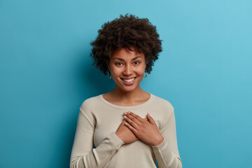 Kind positive African American woman expresses gratitude, has good feeling, presses hands to heart, smiles pleasantly, appreciates someones help, dressed in caual clothes, isolated on blue background
