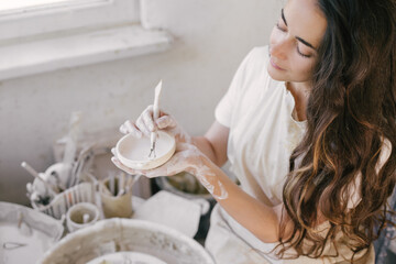 Young beautiful woman with long curly hair in white apron creating handmade ceramic bowl in a pottery.