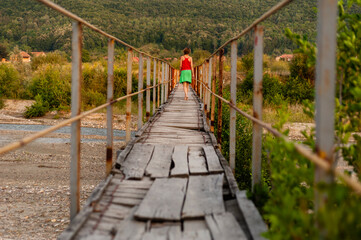 Unrecognizable girl with her back turned walking on a pedestrian bridge in the countryside wearing a red shirt and a green skirt.