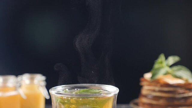 Flavored tea is steaming on black background with honey and stack of pancakes in disfocus. Front Shot. close-up. steam rising above cup with herbal tea. Slow motion. Full hd