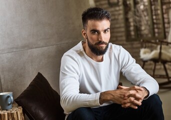 Fototapeta premium Portrait of trendy bearded man in casual clothes sitting at home, looking at camera. 