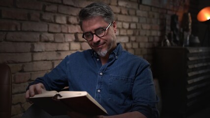 Mature man in his 50s reading book at home sitting in dark living room. 