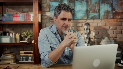 Mature man working from home, sitting at desk in home office using laptop, thinking, browsing internet, teleworking online. 