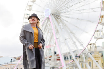 Young woman wearing hat walking outdoors on the city street near ferris wheel smiling cheerful.