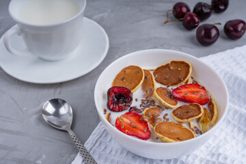 Breakfast consisting of a bowl full of mini pancakes, strawberry, cherry slices and milk laying on white napkin near spoon and cherries together with cup of milk on concrete surface