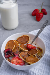 White bowl full of mini pancakes, strawberry and cherry slices and milk together with silver spoon laying on white napkin against jar with milk on concrete surface at kitchen