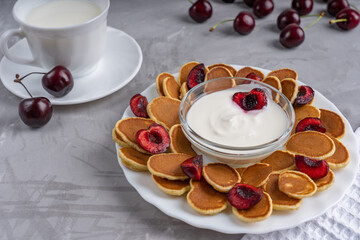 Plate with mini pancakes and cherry slices and a bowl of white yogurt together with cup of milk are laying on concrete surface at kitchen