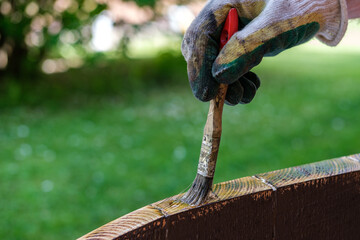 A man (painter) paints the ends of a wooden tabletop with varnish, in the background you can see a garden and a lawn with green grass.