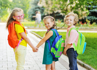 Fototapeta na wymiar three school children-two girls and a curly-haired boy with school colored bags smiling hold hands and go to school. Back to school concept, September 1, knowledge day, education.
