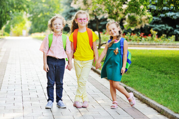 three school children-two girls and a curly-haired boy with school colored bags smiling hold hands and go to school. Back to school concept, September 1, knowledge day, education.