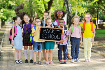 a group of multi-racial school children in colorful clothes, carrying school bags and backpacks hold a sign that reads "Back to school" against the background of greenery and a Park.