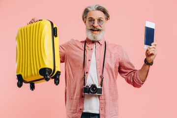 Stylish bearded man with a camera holds a yellow suitcase and a passport with a ticket, smiles and prepares for the trip.