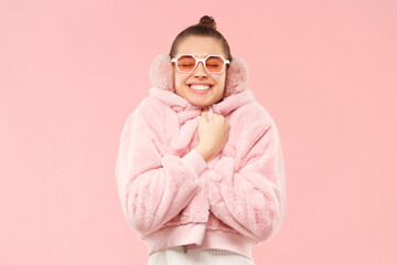 Young happy girl in cozy fur coat and colored glasses, wrapping herself to feel warm, enjoying her winter holidays, isolated on pink background