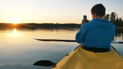 man takes a photo of the sunset on a smartphone on the lake sitting in a kayak with a paddle, self-isolation outdoor activities alone