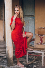 Stylish and elegant woman in a red dress is resting near the house. posing with a cigarette near the column.