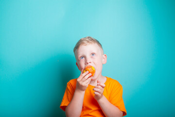 Cute and funny baby boy in an orange t-shirt bites off an orange juicy peach. The child is a blond European on a blue background. Health benefits of fresh fruit.